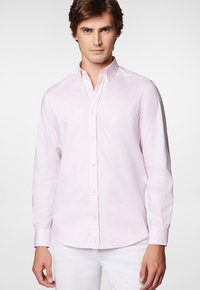 Jasnoróżowa koszula z długim rękawem na guziki, uszyta z gładkiego materiału, z kołnierzykiem w szpic i białymi guzikami, noszona z białymi spodniami.