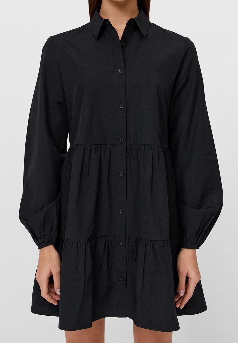 Robe noire à boutons sur le devant, avec un col, des manches bouffantes longues et une jupe à volants. Fabriquée en tissu doux au toucher lisse.