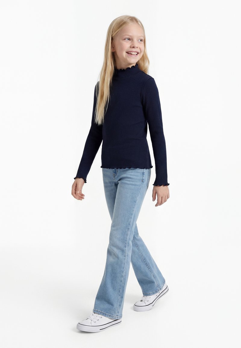 Junges Mädchen läuft, trägt ein marineblaues geripptes Langarmshirt, hellblaue Jeans und weiße Sneakers, lächelt und schaut seitwärts.