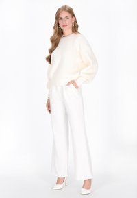 Maglione bianco a maglia con texture a costine, abbinato a pantaloni bianchi a gamba larga e tacchi a spillo bianchi. La modella posa con una mano in tasca.