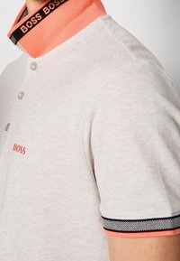 Jasno beżowa koszulka polo z koralowym kołnierzykiem, czarnym i białym wykończeniem, z logo "BOSS" na kołnierzyku i na klatce piersiowej, noszona na górnej części ciała.