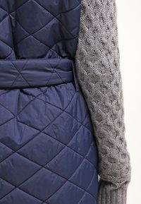 Marinblå kviltad puffer-väst med belagd midja och grå texturerade stickade ärmar med ett mönster av upphöjda cirklar. Mjuk tygtextur.