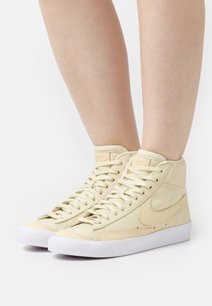 Wysokie sneakersy w kremowym zamszu z białą gumową podeszwą, z teksturowanym wykończeniem i wyraźnym logo po bokach. Sznurowadła przewleczone.