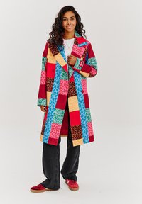 Farebný patchworkový kabát s červenými, modrými, ružovými a žltými štvorcami so zvieracími vzormi. Vyrobený z látky s pevnou, nadrozmernou konštrukciou.