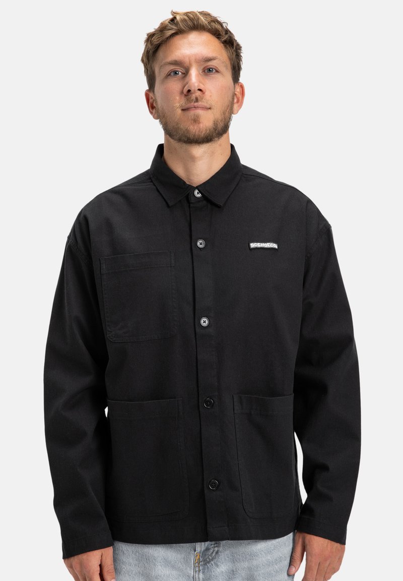 Czarna kurtka typu button-up z solidnego materiału, z kołnierzykiem, dwoma przednimi kieszeniami i małą etykietą z logo na piersi.