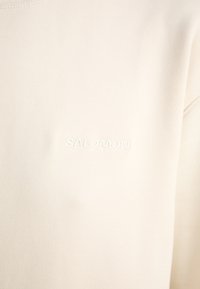 Sweter w kolorze kremowym z teksturowanej tkaniny oraz subtelnym haftowanym logo "Salomon". Klasyczny okrągły dekolt i luźny krój.