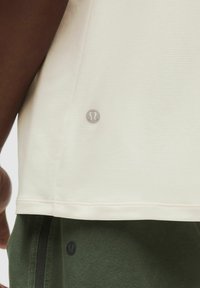 Svetlo bež kratkolasna športna majica z teksturirano tkanino in srebrnim logotipom na robu, v kombinaciji s temno zelenimi hlačami z detajlom logotipa.