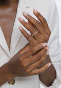 Tynd, pastelgrøn ring med blomsterdetaljer og to små lyserøde sten, båret på en hånd med velplejede, naturlige negle.