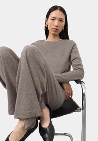 Donna con lunghi capelli neri, che indossa un maglione di maglia taupe e pantaloni larghi, orecchini d'oro e ballerine nere, seduta su una sedia cromata e nera.