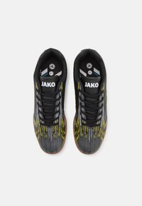 Des chaussures de sport noires avec une tige texturée, présentant des accents jaunes et gris, des lacets noirs et des semelles en caoutchouc brun. Logo "JAKO" sur la languette.