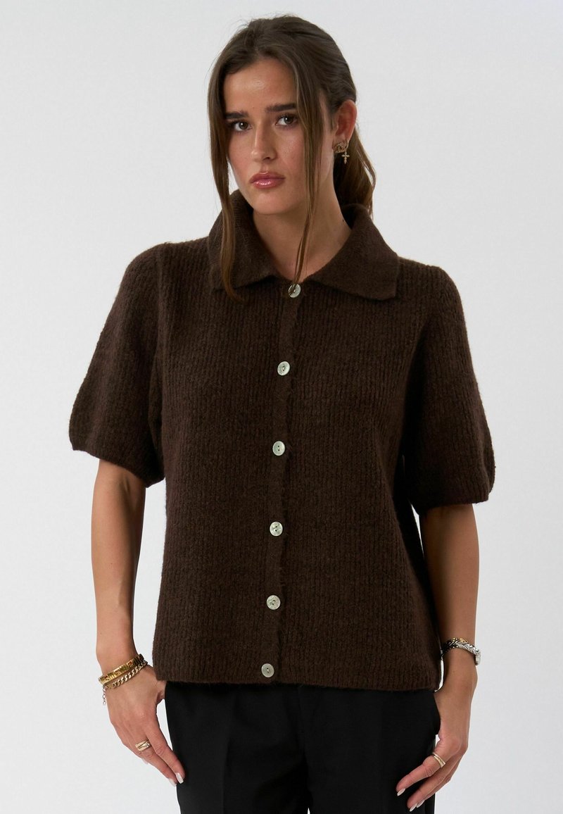 Jeune femme portant un cardigan tricoté à manches courtes marron foncé avec des boutons, un pantalon noir et des bracelets dorés au poignet.