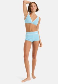 Kvinne står barbeint med en arm hevet, iført en lys blå teksturert bikini topp og matchende høy midje shorts med hvit kant.