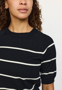 Črni pulover iz pletenega materiala s belimi vodoravnimi črtami, kratkimi nabranimi rokavi in okroglim izrezom. Gladek teksturni videz in oprijet kroj.