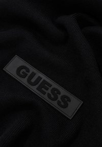 Tessuto a maglia nera con un patch in gomma texturizzato con il logo "GUESS". Il tessuto è liscio, presenta una leggera costina e un aspetto morbido.