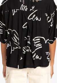 Camicia nera oversize con motivi astratti bianchi. Tessuto morbido, maniche corte e scollatura rotonda con perline decorative nella parte superiore.