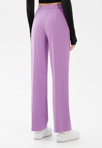 Kalhoty s vysokým pasem a širokými nohavicemi v světle fialovém materiálu s hladkou texturou. Mají přizpůsobený střih a poutka na opasek, v kombinaci s bílými teniskami.