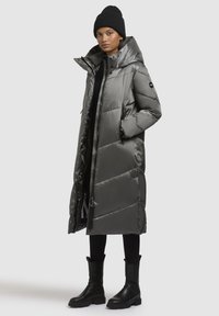 Grå, lång pufferjacka med en glänsande yta, featuring en hög krage, dragkedja fram och sidofickor. Bärs med svarta byxor och ankelboots.