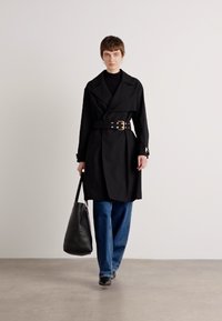 Černý trench coat s páskem a širokými klopami, spárovaný s černým topem a volnými modrými džíny. Modelka nese velkou černou tašku. Hladká textura.