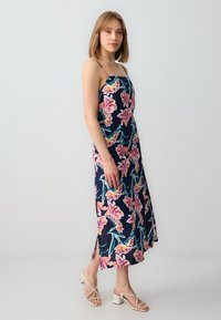 Marineblaues Maxikleid mit pinken und grünen Mustern, dünnen Trägern und seitlichen Schlitzen. Kombiniert mit weißen High-Heel-Sandalen.