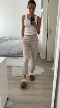 Smėlio spalvos pritaikyti kelniai su švariu kirpimu, derinami su rudo odos šlepetėmis, turinčiomis baltą pūkinę apdailą. Baltas tank top'as užbaigia įvaizdį.