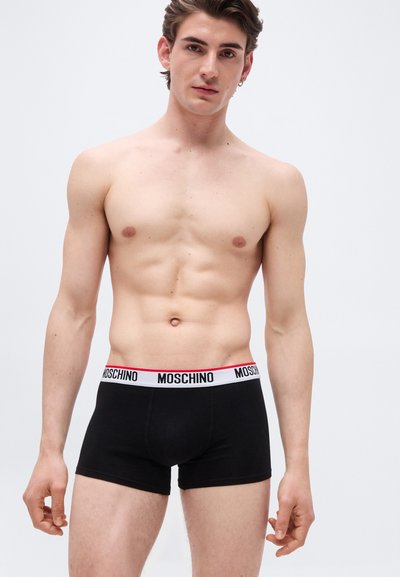 Νεαρός άνδρας χωρίς μπλούζα, φορώντας μαύρα βρακάκια Moschino με λευκή και κόκκινη λωρίδα με λογότυπο, στέκεται μπροστά σε ένα απλό φωτεινό φόντο.