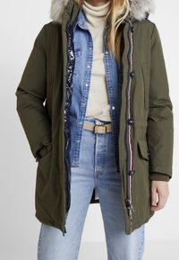 Olivově zelená parka s kapucí lemovanou kožešinou, zapínáním na zip, předními kapsami a pruhovaným detailem. Nositelná přes džínovou košili a béžový svetr.