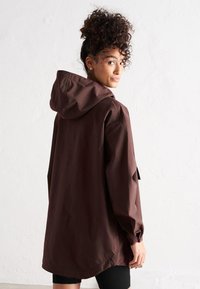 Chaqueta marrón impermeable con capucha, que presenta un corte holgado, mangas largas y una textura suave y ligera.