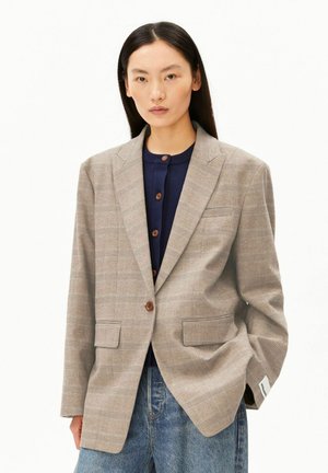 Femme portant un blazer à carreaux beige sur un pull bleu marine boutonné et un jean bleu, debout avec une main dans la poche devant un fond blanc.
