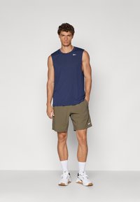 Granatowy, bezrękawnik sportowy połączony z oliwkowymi szortami. Model ma na sobie białe sneakersy z ciemnymi akcentami oraz białe skarpetki. Minimalne logo widoczne.