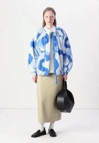 Casaco bomber azul e branco com padrão, com punhos elásticos, combinado com uma saia bege e uma bolsa preta. Estilizado com meias brancas e sapatos pretos.