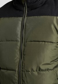 Padded jacket z ciemnozielonym korpusem i czarną, teksturowaną górną częścią. Posiada zamek błyskawiczny z przodu oraz pikowane detale.