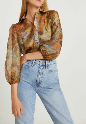 Durchsichtige, mehrfarbige Bluse mit Blumenmuster, Knopfleiste vorne und Puffärmeln; kombiniert mit hoch taillierten hellblauen Jeans.