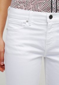 Bílé denimové džíny s klasickým pěti-kapsovým designem, obsahující poutka na pás, jednořadové zapínání na knoflík a kontrastní prošívání.