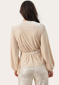 Top de terciopelo beige con mangas largas y diseño envolvente. La tela tiene una textura suave, ceñido en la cintura con un cinturón a juego.