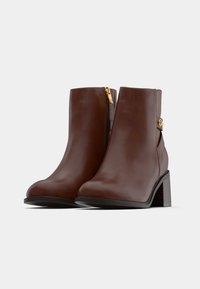 Botine din piele maro, cu o textură netedă, vârf rotunjit, fermoare laterale și toc gros. Prezintă un accent aurie pe lateral.