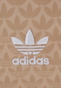 Světle hnědý textil s jemným vzorem listů a bílým vyšitým logem Adidas, včetně tří proužků nad názvem značky.