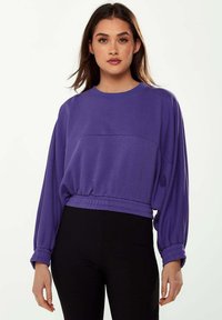 Purple cropped sweatshirt with long sleeves, elastic waistband, and a smooth texture, paired with black ribbed leggings.