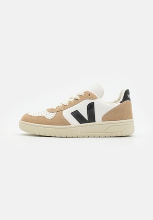 Low-top sneaker with beige suede overlays, white leather panels, cream sole, black "V" logo on side, and white laces on a white background.
