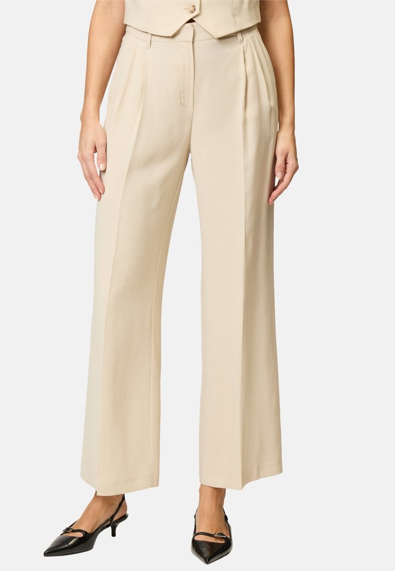 Pantalon beige taille haute à jambes larges avec plis, porté avec des escarpins slingback noirs à bout pointu.