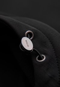 Tela negra con un ajuste metálico y ojetes. Presenta una textura suave y un cordón elástico para un ajuste seguro. Presencia de marca.