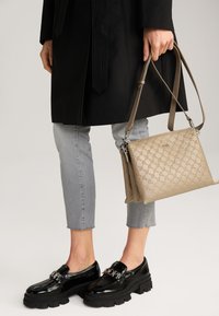 Mocassini neri in vernice con suola spessa, jeans grigi a vita corta e una borsa beige con motivo e tracolla.
