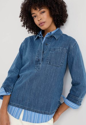 Donkerblauwe denimshirt met een knoopsluiting, een patchzak en gelaagde lichtblauwe gestreepte manchetten die zichtbaar zijn bij de mouwen.