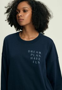 Granatowy sweter z okrągłym dekoltem i długimi rękawami, na przedzie z białym napisem: "MARZ, PLANUJ, CIESZ SIĘ." w pionowym układzie.