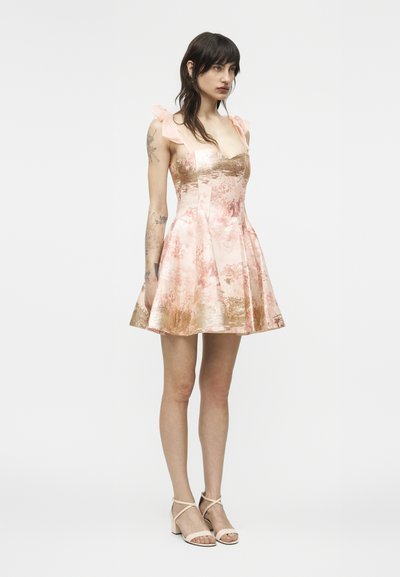 Femeie cu păr închis la culoare, purtând o rochie fără mâneci, în linie A, roz cu modele florale aurii, și sandale bej cu toc, stând în picioare pe un fundal deschis, simplu.