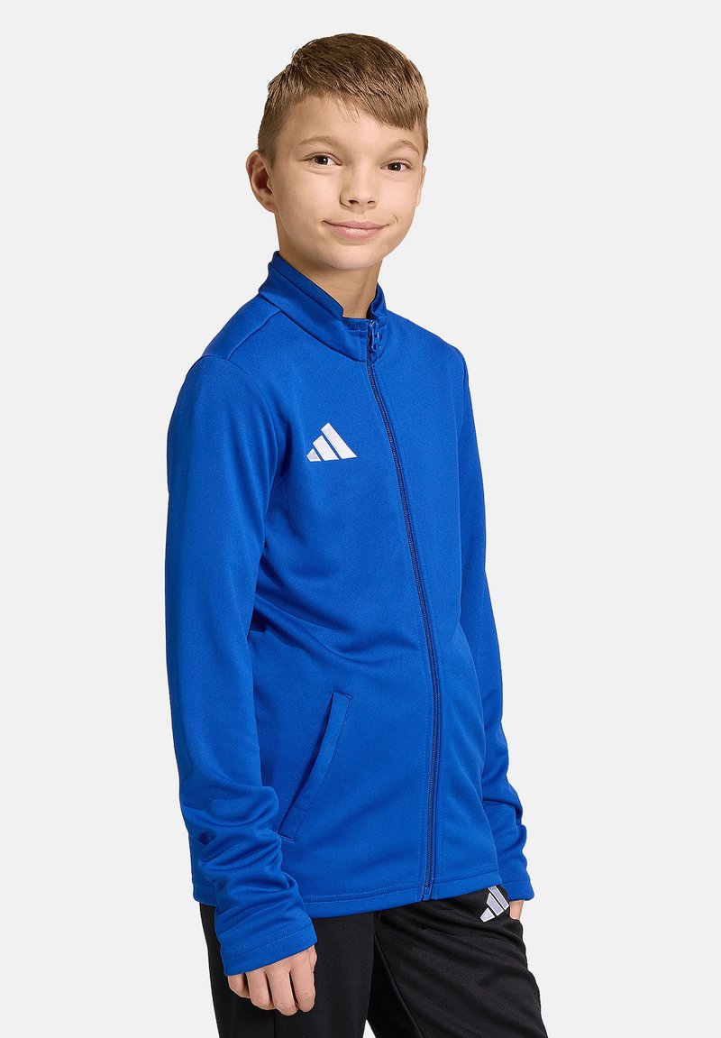 Jeune garçon debout, portant une veste de sport zippée bleue et un pantalon noir avec un logo blanc sur un fond clair uni.