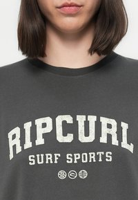 Asmuo, vilkintis tamsiai pilką marškinėlį su baltais užrašais "RIP CURL SURF SPORTS" ir trimis mažomis apvaliomis logotipais žemiau užrašo.
