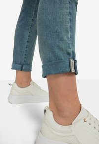 Ljust blå denimjeans med uppvikt fåll, med en etikett på fållen. Matchat med vita sneakers som har en texturerad yta och tjocka sulor.