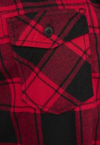 Tessuto flanella a quadri rosso e nero con una distintiva tasca a petto quadrata, caratterizzata da un singolo bottone nero e dettagli di cucitura visibili.
