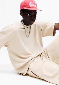 Béžové oversized tričko a odpovídající šortky, vyrobené z hladkého materiálu. Model má na sobě růžovou čepici s bílým textem a vícevrstvé stříbrné náhrdelníky.
