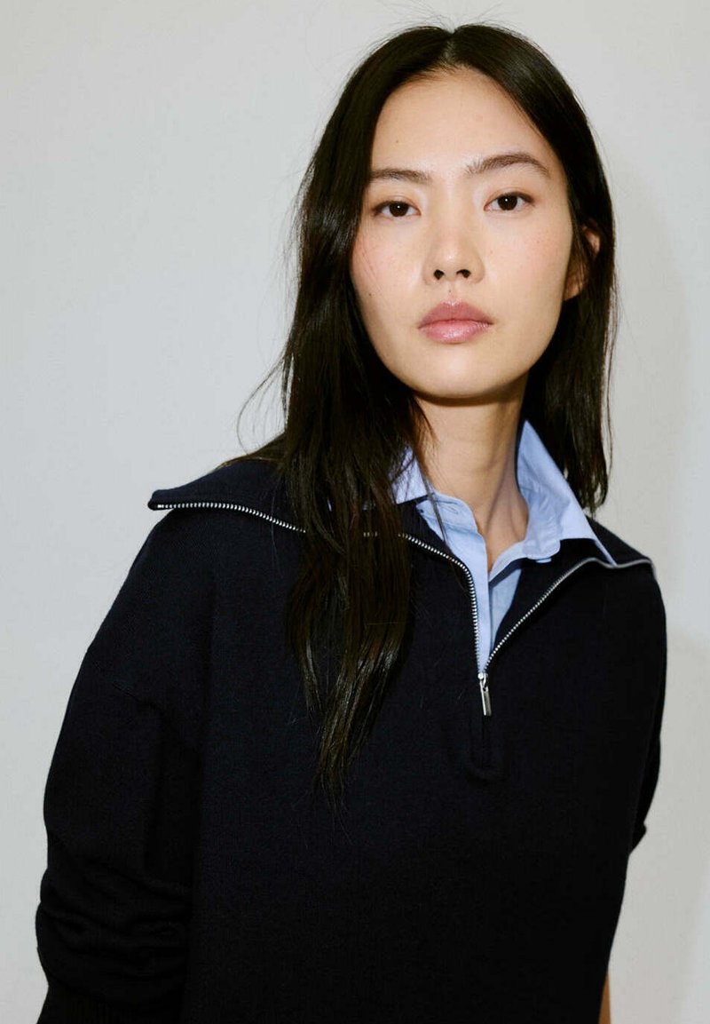 Jeune femme aux longs cheveux noirs portant un pull zippé noir par-dessus une chemise à col bleu clair sur un fond uni.